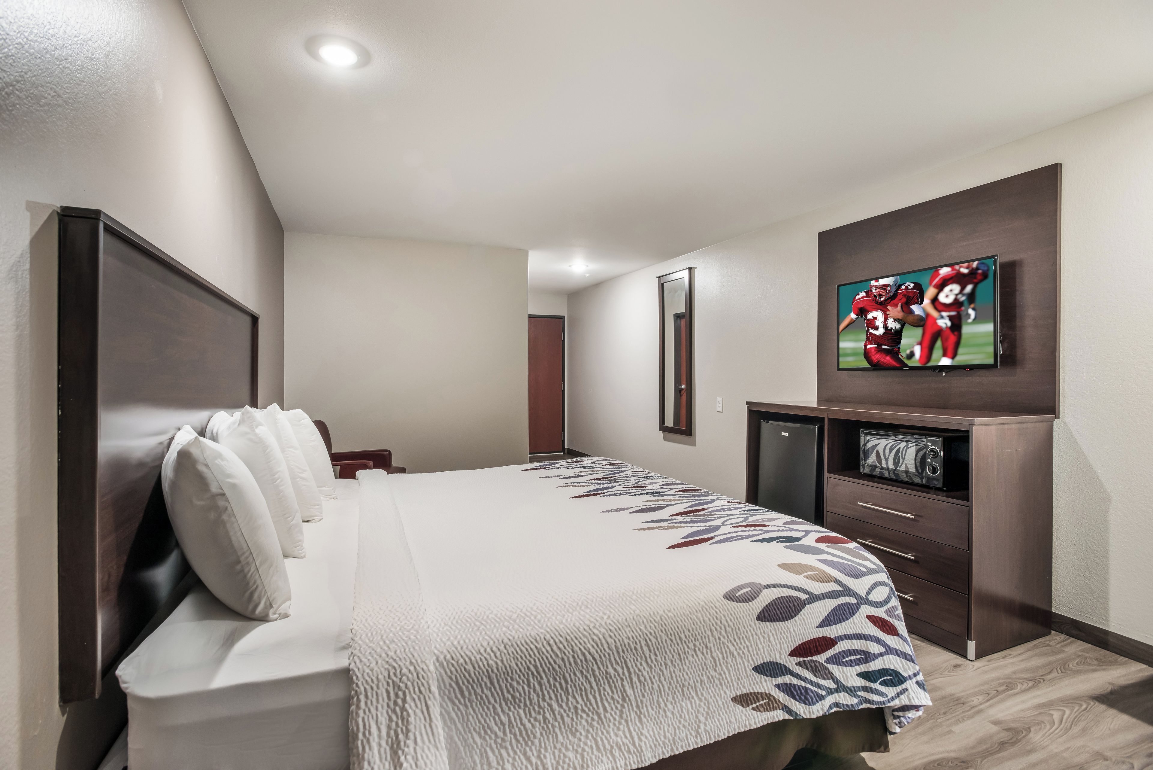 A hotel room with a bed, a television mounted on the wall, a small cabinet, and a door leading to another room.