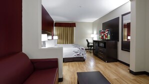 A hotel room with a bed, a desk, a TV, a red sofa, and a wooden floor.