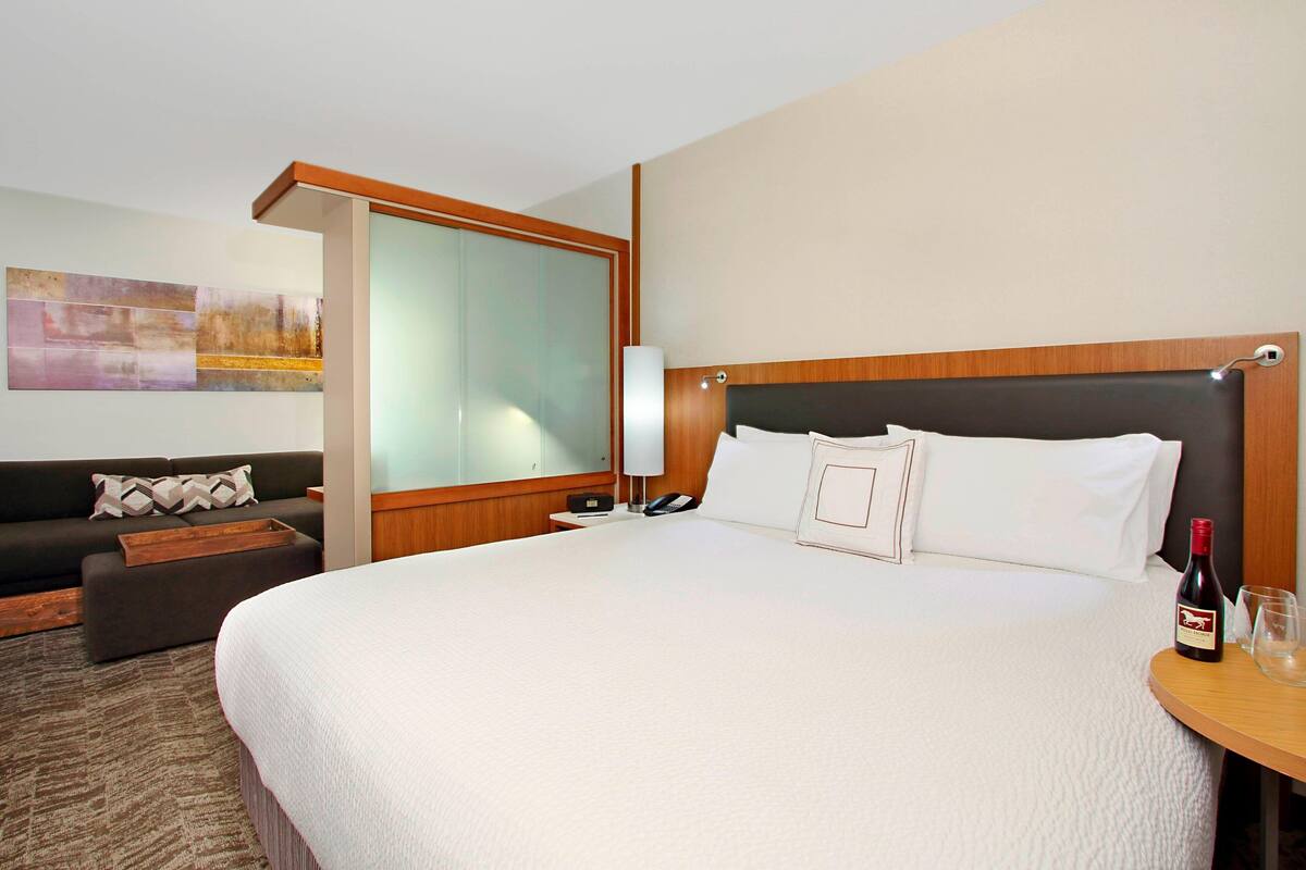 Habitación de hotel con una cama grande, un sofá, una mesita de noche con una botella de vino y una copa, y un espejo en la pared.