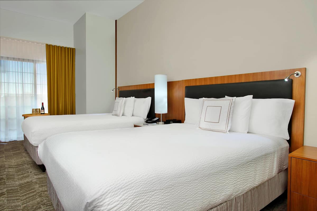 Habitación de hotel con dos camas, cabecero de madera, mesita de noche con lámpara y ventana con cortinas.