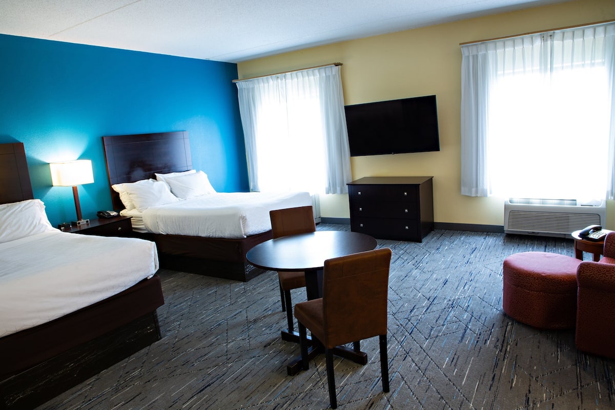 A hotel room with two beds, a dining table with chairs, a TV mounted on the wall, and a window with curtains.