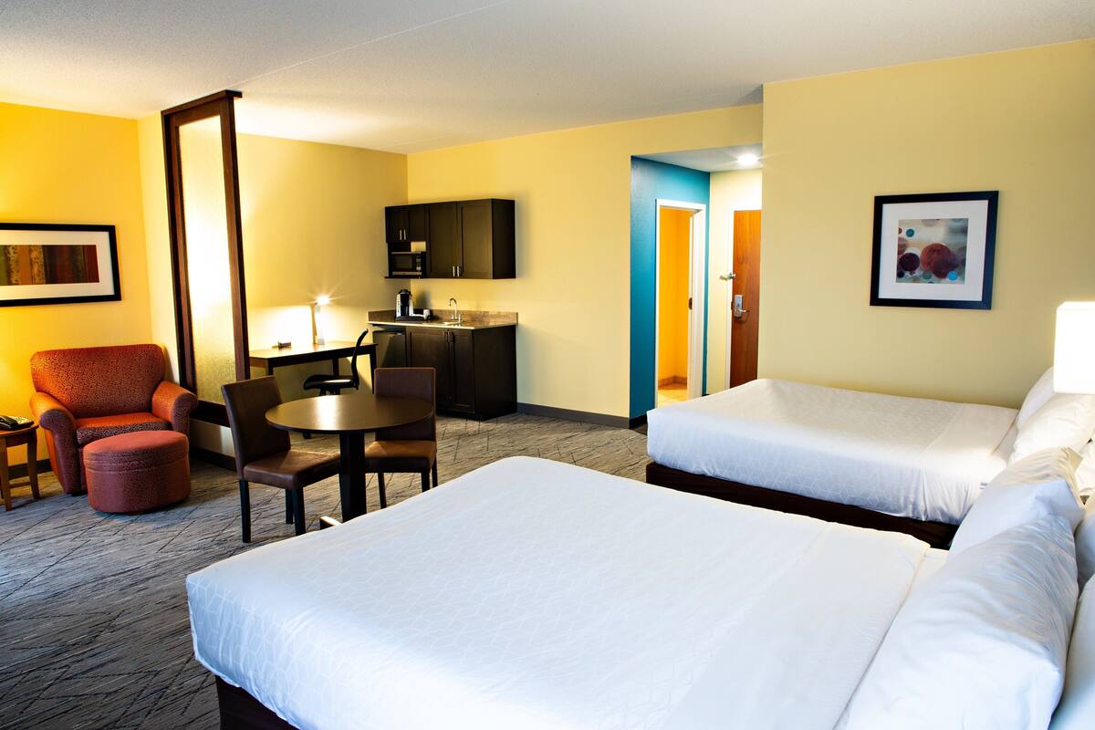 A hotel room with two beds, a desk, a chair, a small table, and a kitchenette.