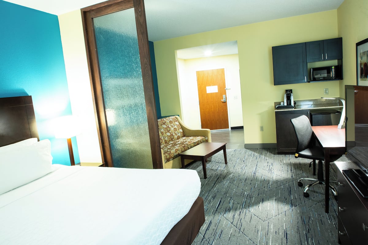 A hotel room with a bed, a sofa, a desk, a chair, a microwave, and a small kitchenette.