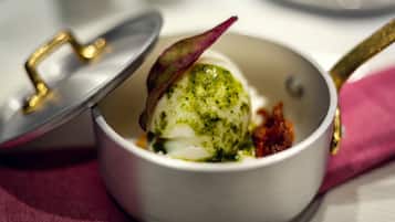 A white bowl with a lid, containing a white spherical food item, possibly a dessert, with a green topping and a purple leaf garnish.