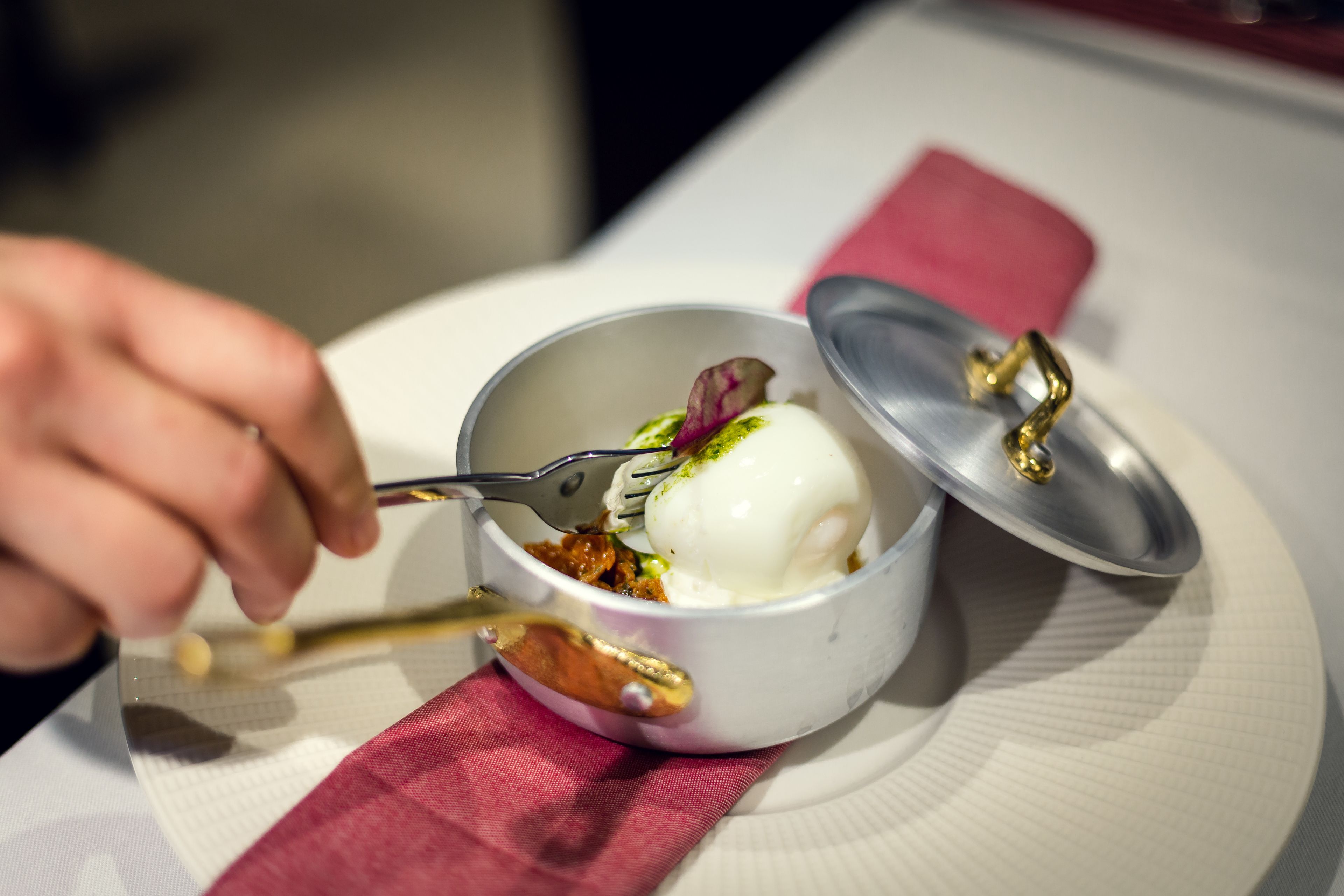 A dessert in a white bowl with a lid, a spoon, and a napkin on a table.