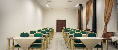 Un salón de conferencias con filas de sillas verdes y mesas, una puerta de madera al fondo y una ventana con cortinas.