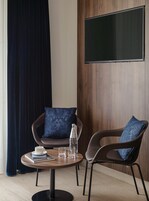 Quarto moderno com uma mesa de madeira redonda, duas cadeiras, televisão de tela plana fixada em uma parede de madeira e uma janela grande com cortinas escuras.