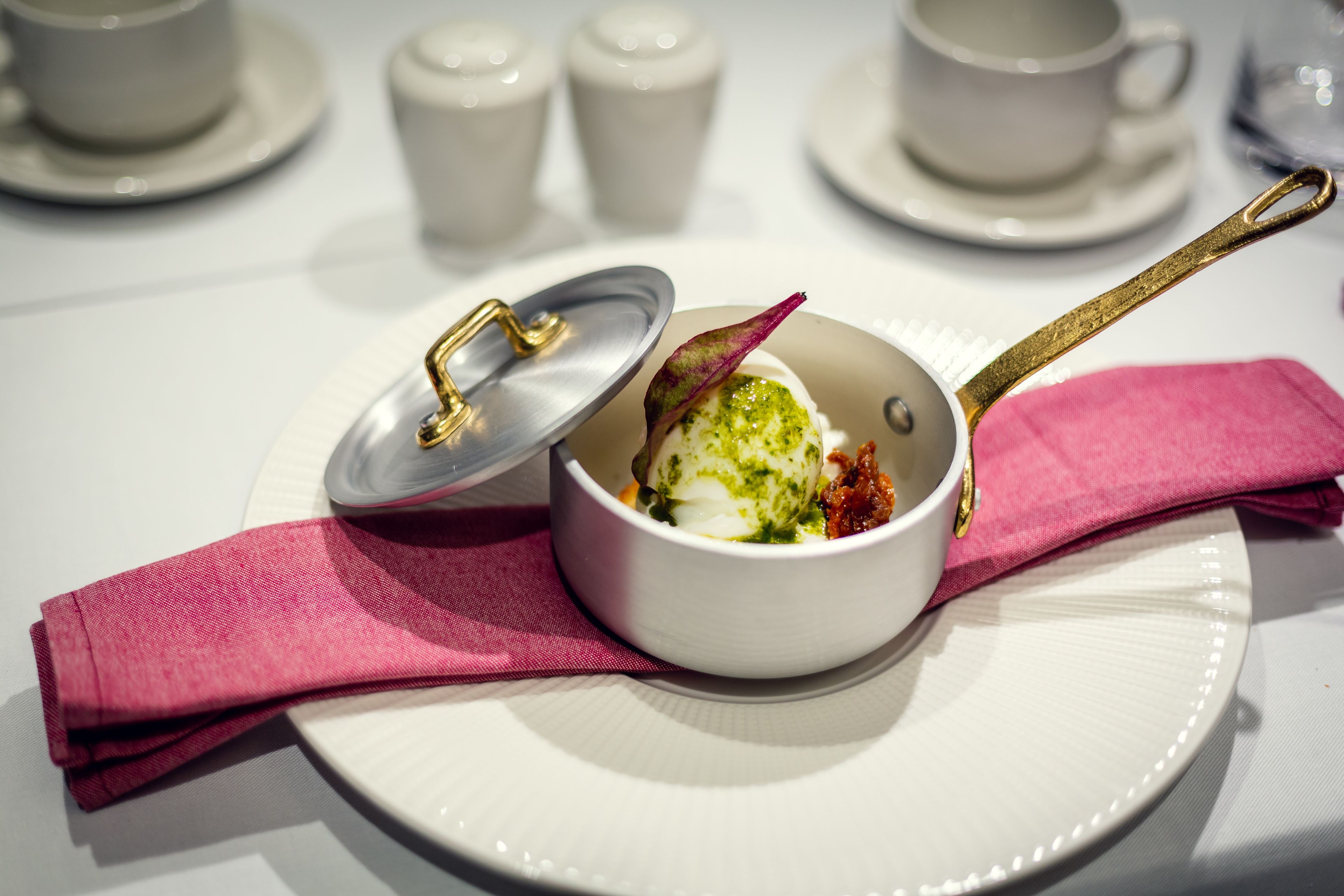 A white bowl with a lid on a white plate, accompanied by a pink napkin and a gold spoon.