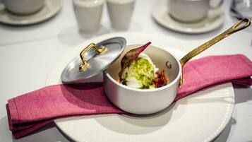 A white bowl with a lid on a white plate, accompanied by a pink napkin and a gold spoon.