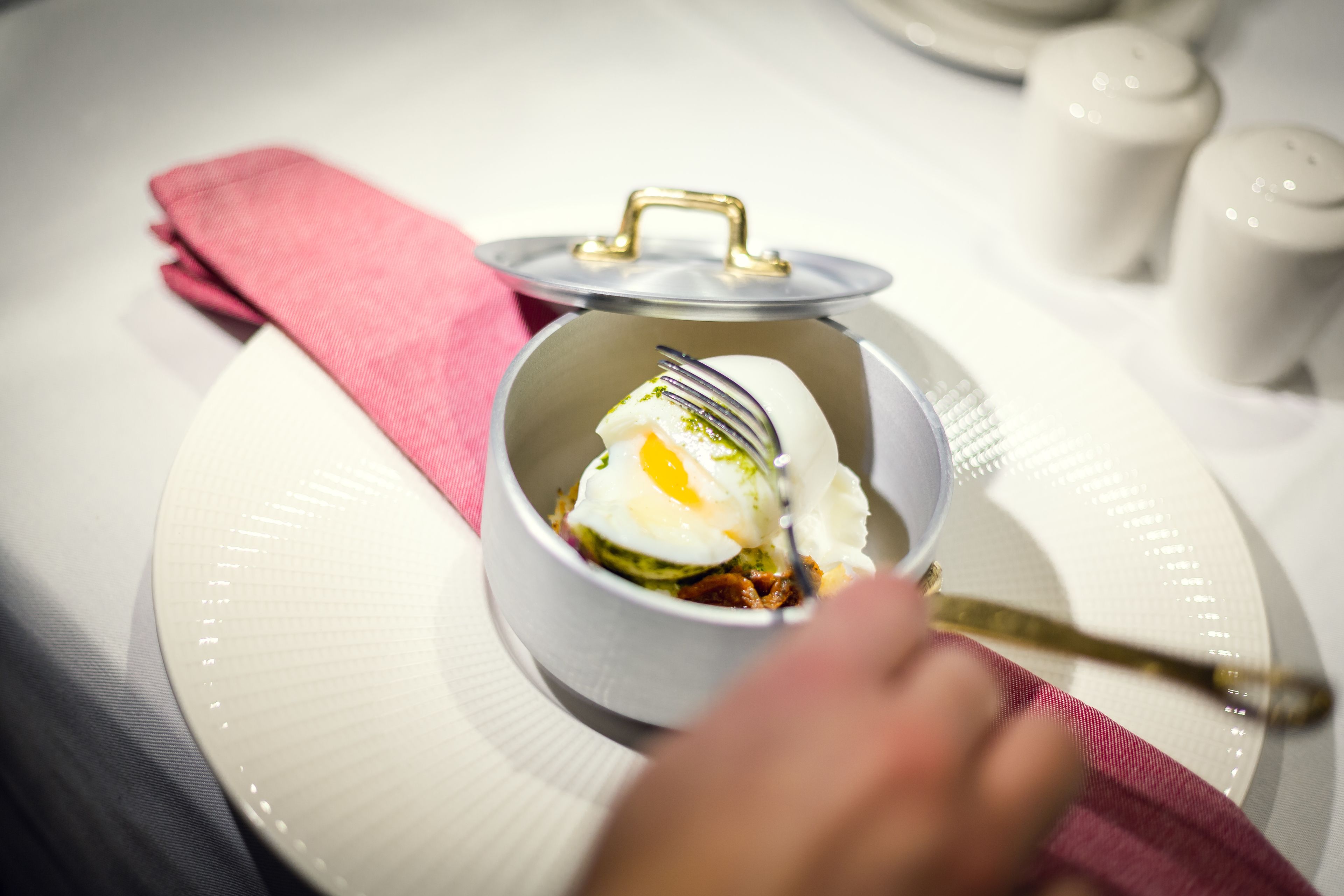 Un tazón blanco con tapa sobre un plato blanco, un tenedor y una servilleta rosa.