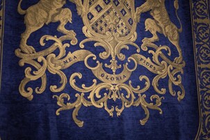 Primeiro plano de um tecido azul com bordado dourado exibindo as palavras “HEA”, “FIDES” e “GLORIA”.