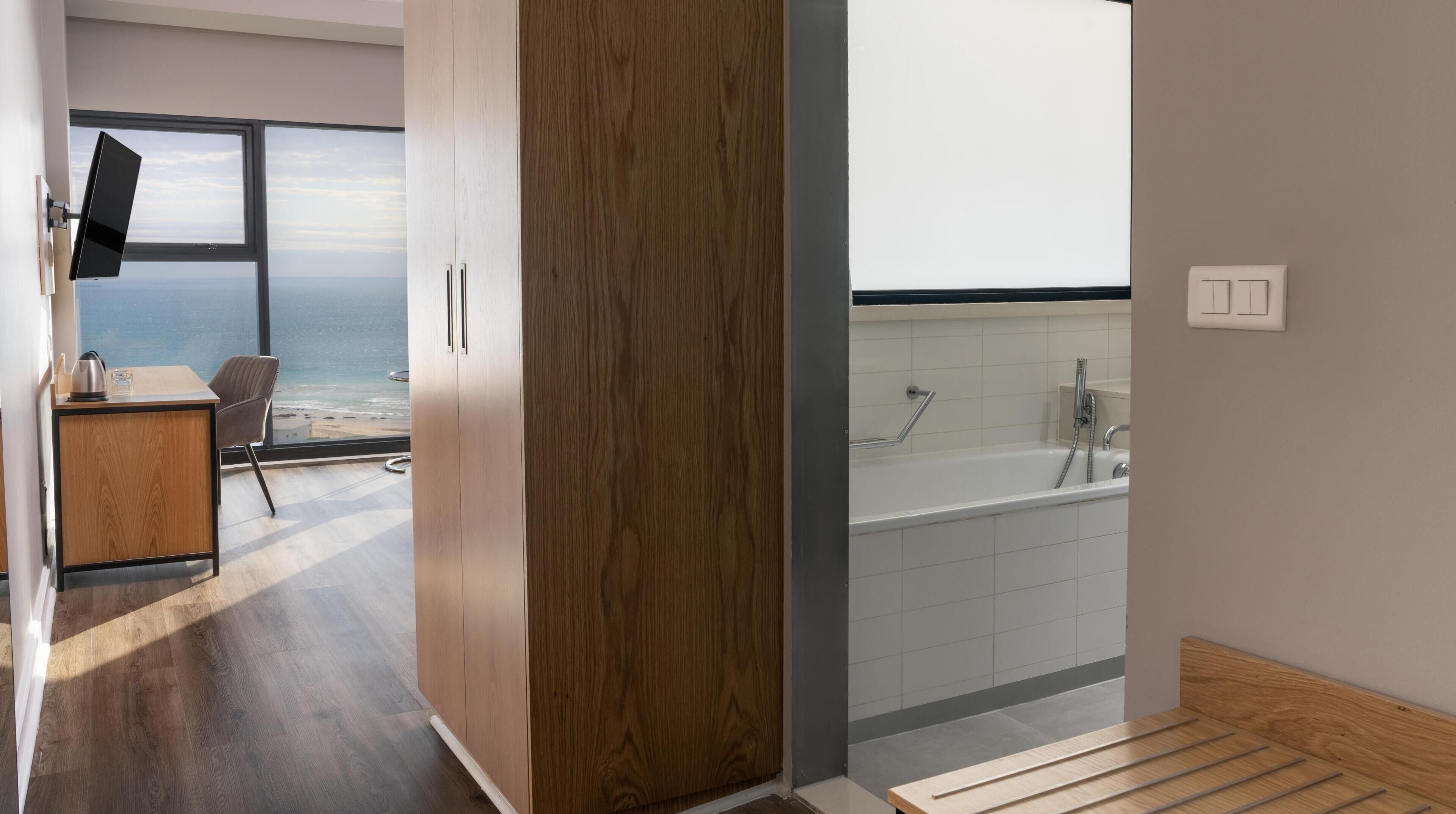 A modern hotel room with a wooden wardrobe, a desk with a kettle, a chair, a large window with a view of the sea, and a bathroom with a bathtub and shower.
