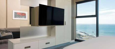 A modern hotel room with a large window offering a sea view, a flat-screen TV mounted on a white cabinet, and a bed with a grey textured rug.
