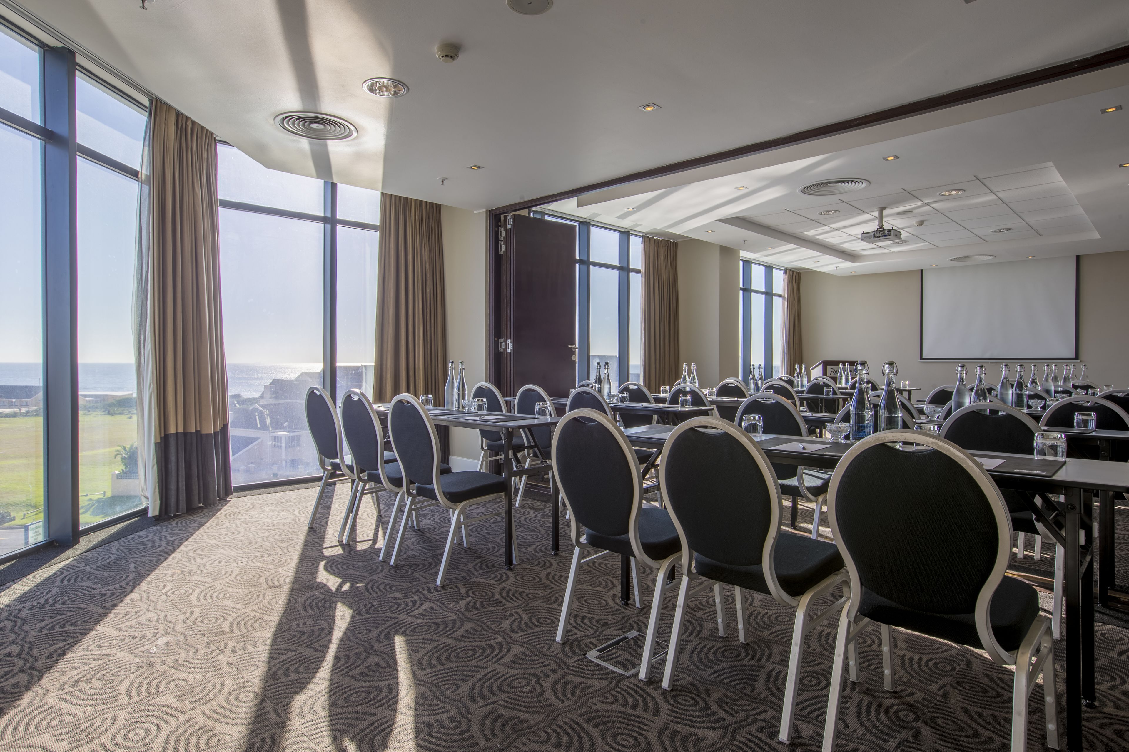 Un salón de conferencias con mesas y sillas, ventanas amplias y una pantalla para proyector.