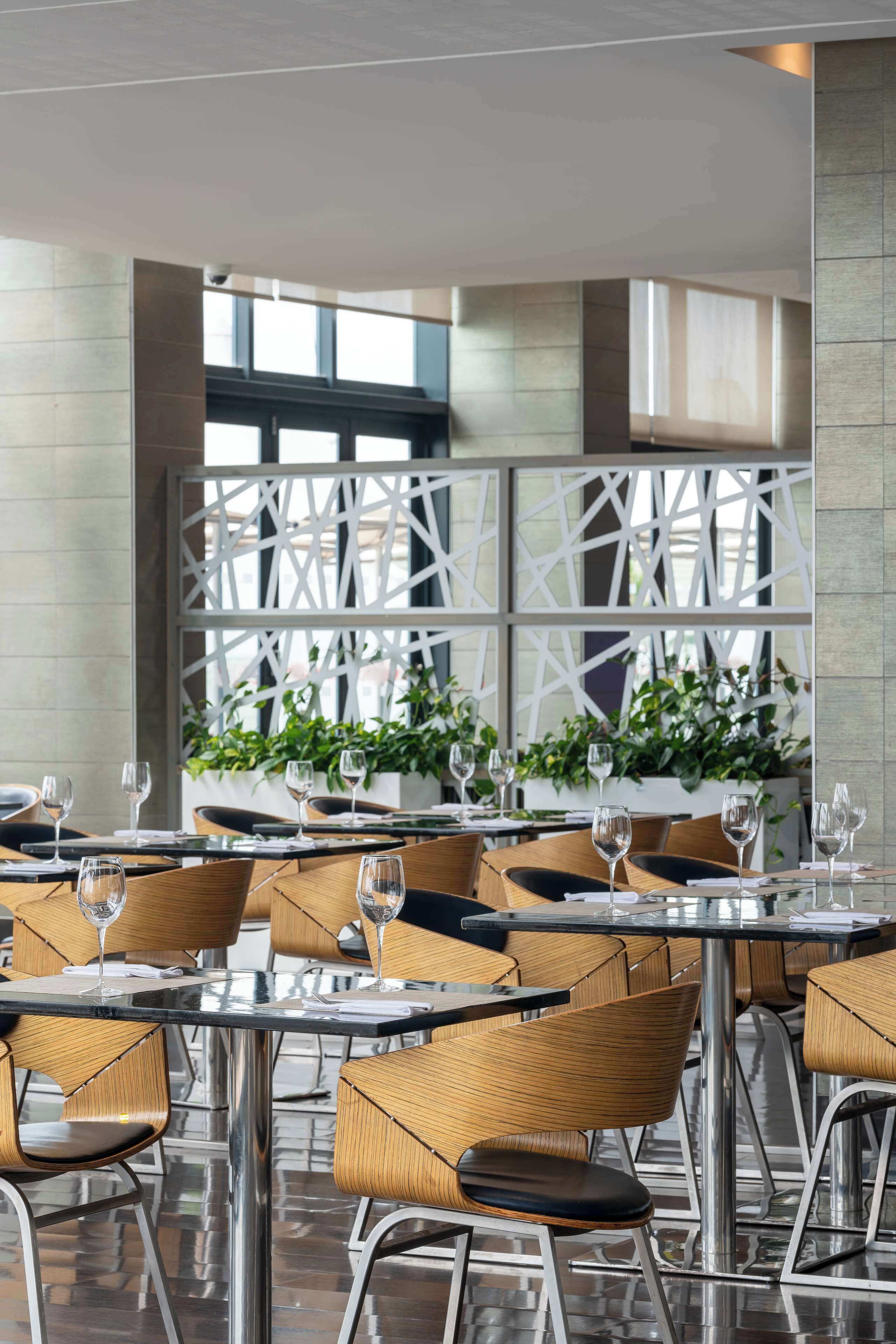 A modern restaurant with glass tables and wicker chairs, featuring a geometric white partition and potted plants.