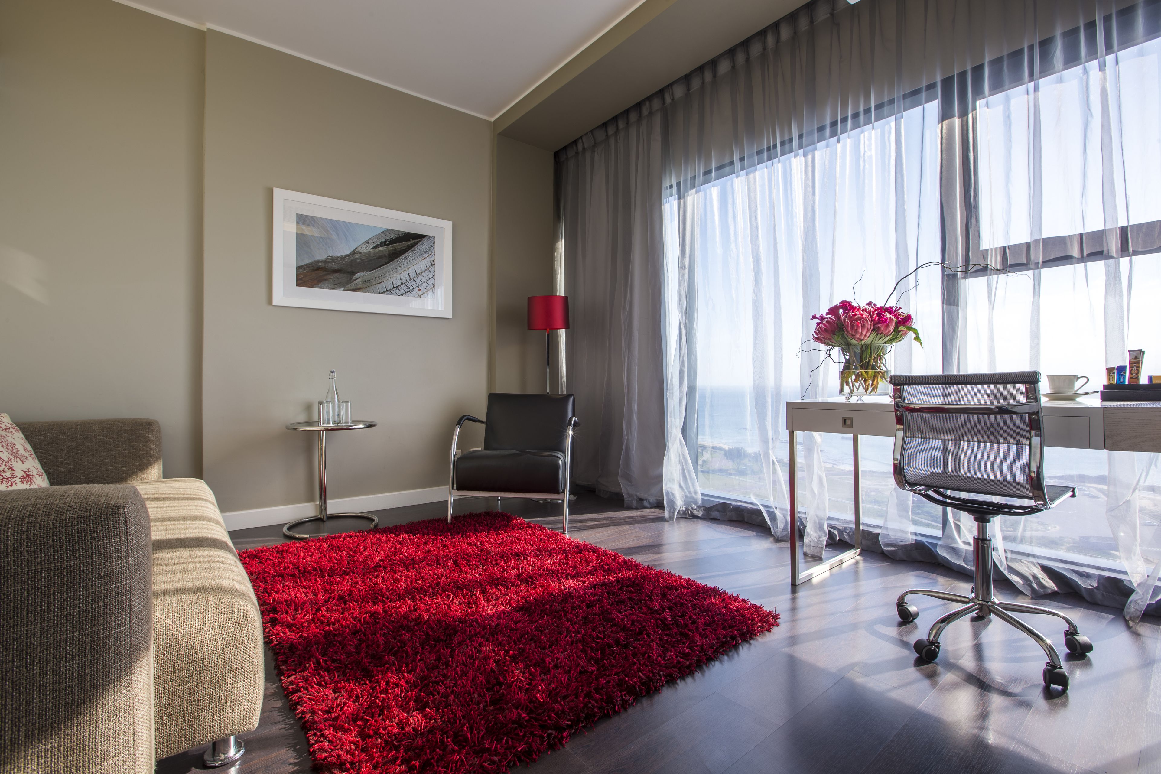 A modern hotel room with a large window, a red rug, a desk with a chair, and a framed picture on the wall.