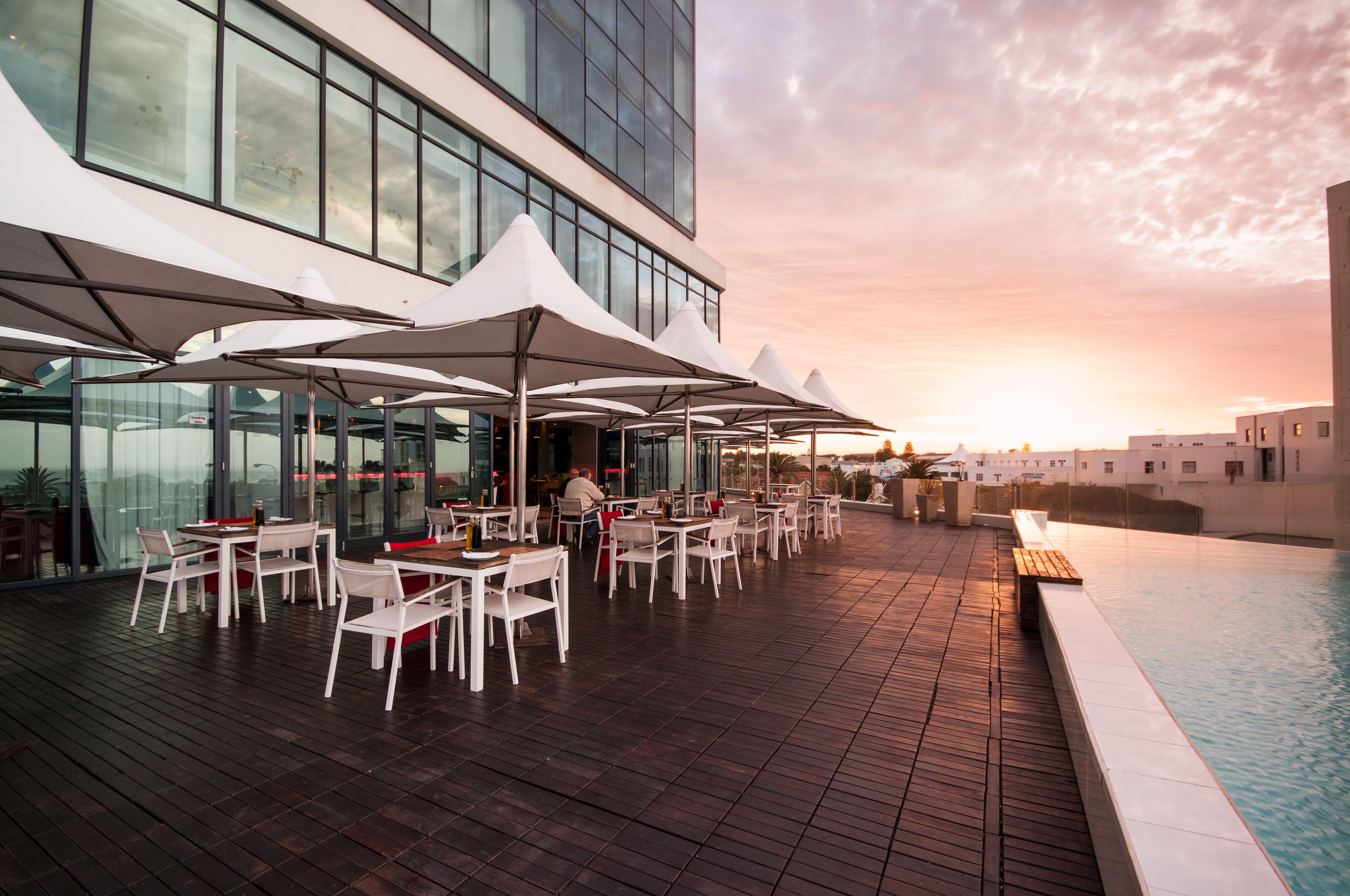 Un área de comedor al aire libre con mesas y sillas, una piscina y un edificio moderno de fondo.