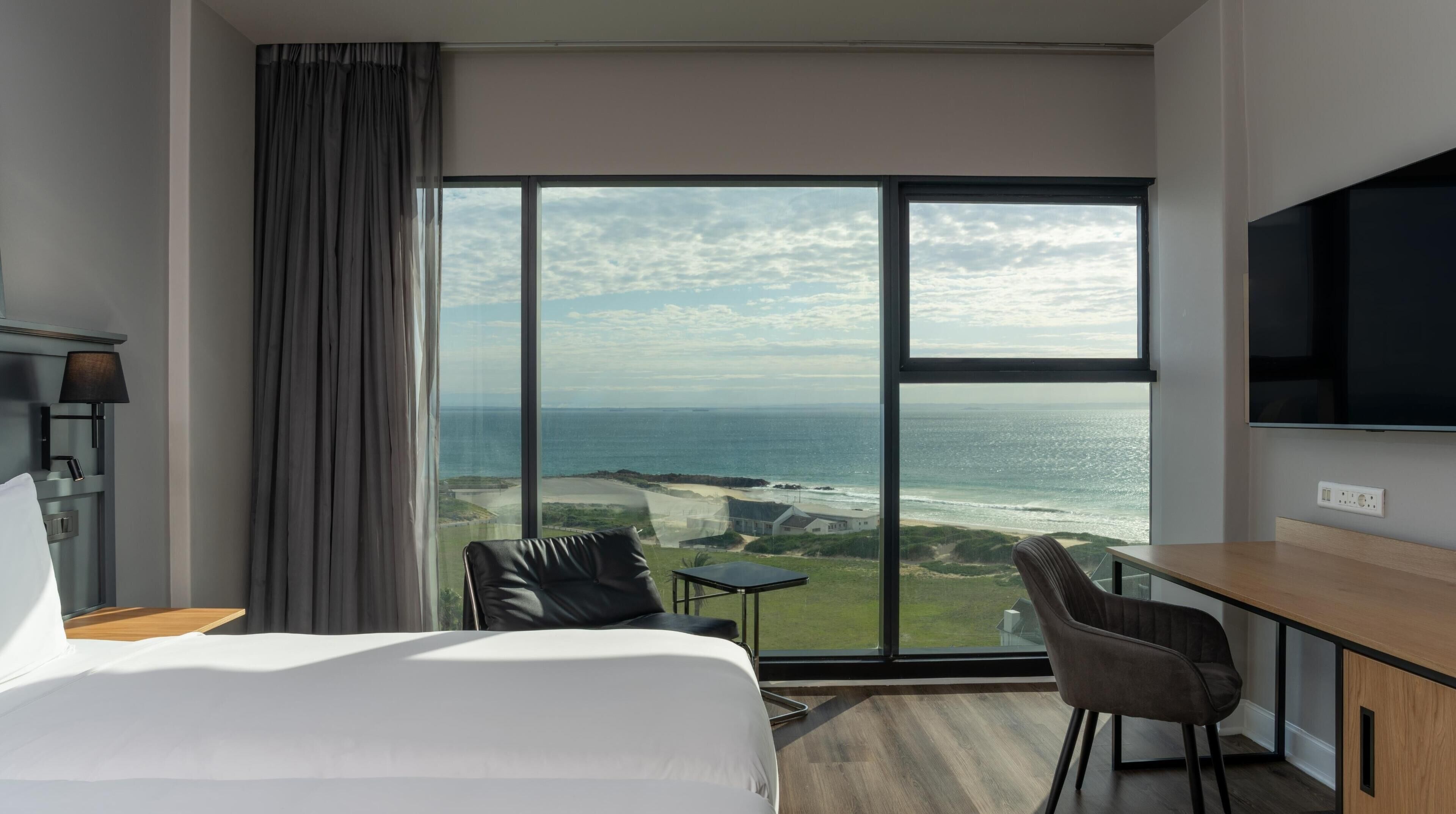A hotel room with a large window offering a view of the ocean and coastline, a bed, a desk with a chair, and a flat-screen TV.