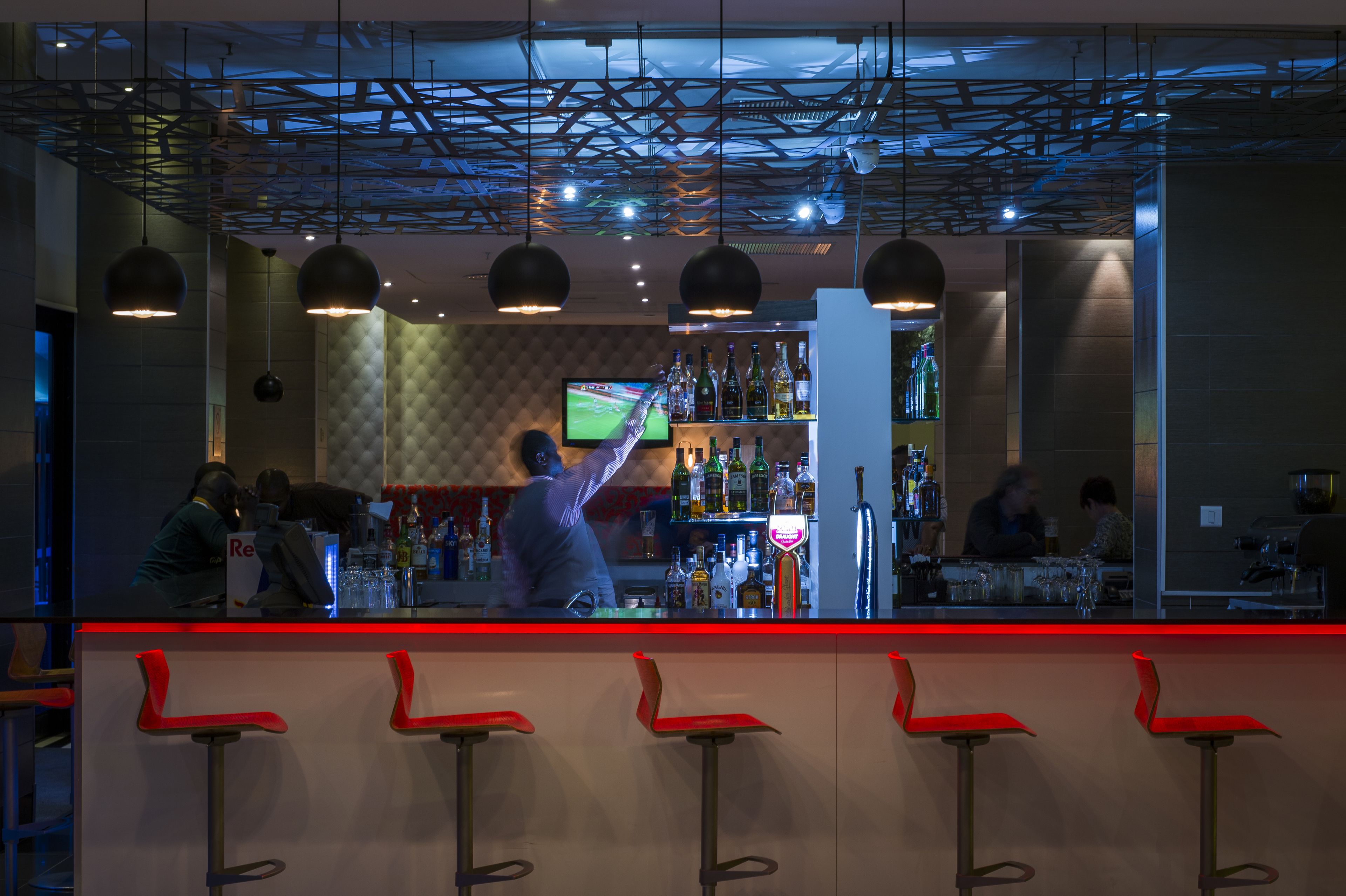 Zona de bar con un cantinero, taburetes rojos y una pantalla de televisión.