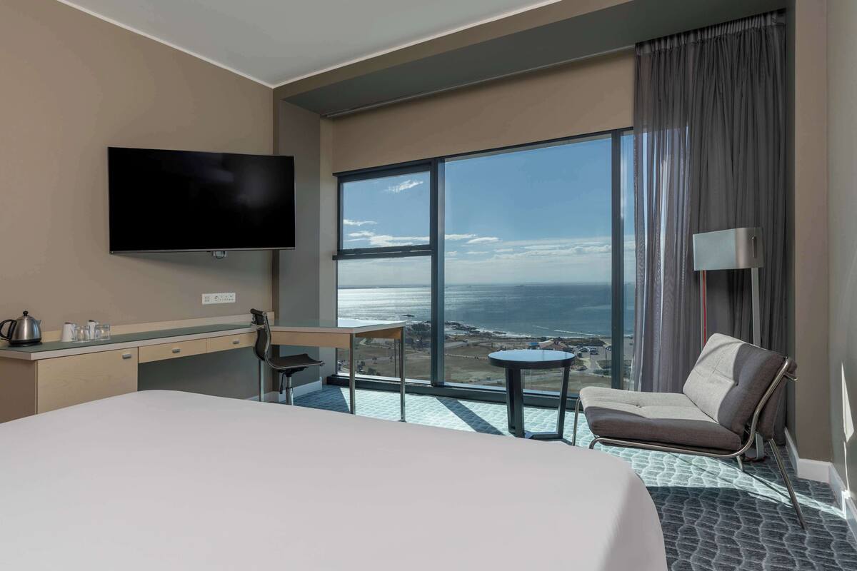 Ein Hotelzimmer mit einem großen Bett, einem Schreibtisch, einem Sessel, einem Fernseher, einem kleinen Tisch und Meerblick.