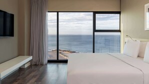 Une chambre d’hôtel dotée d’une grande fenêtre donnant sur la mer, d’un lit, d’une banquette et d’un téléviseur à écran plat.