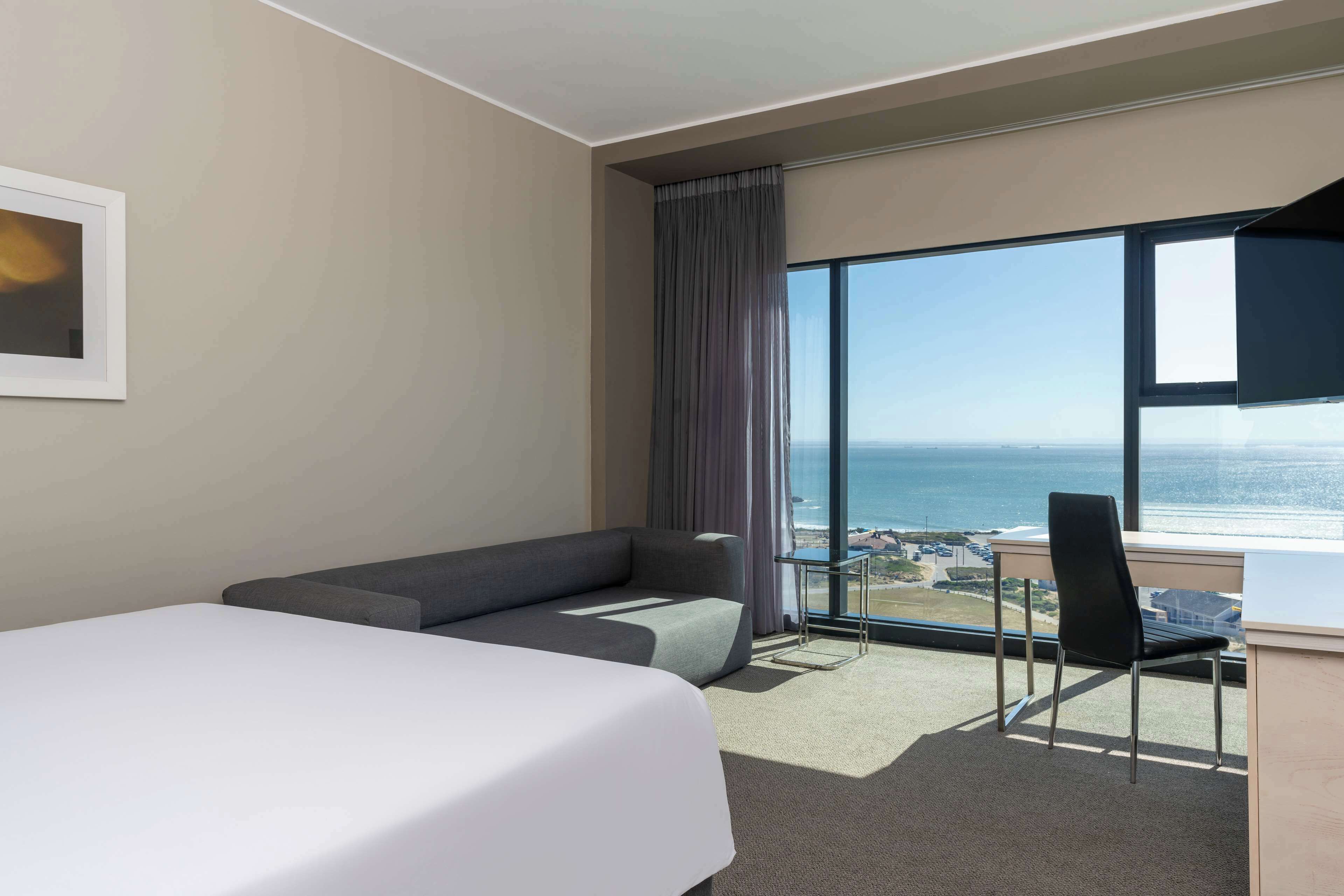 A hotel room with a bed, a sofa, a chair, and a large window with a view of the ocean.