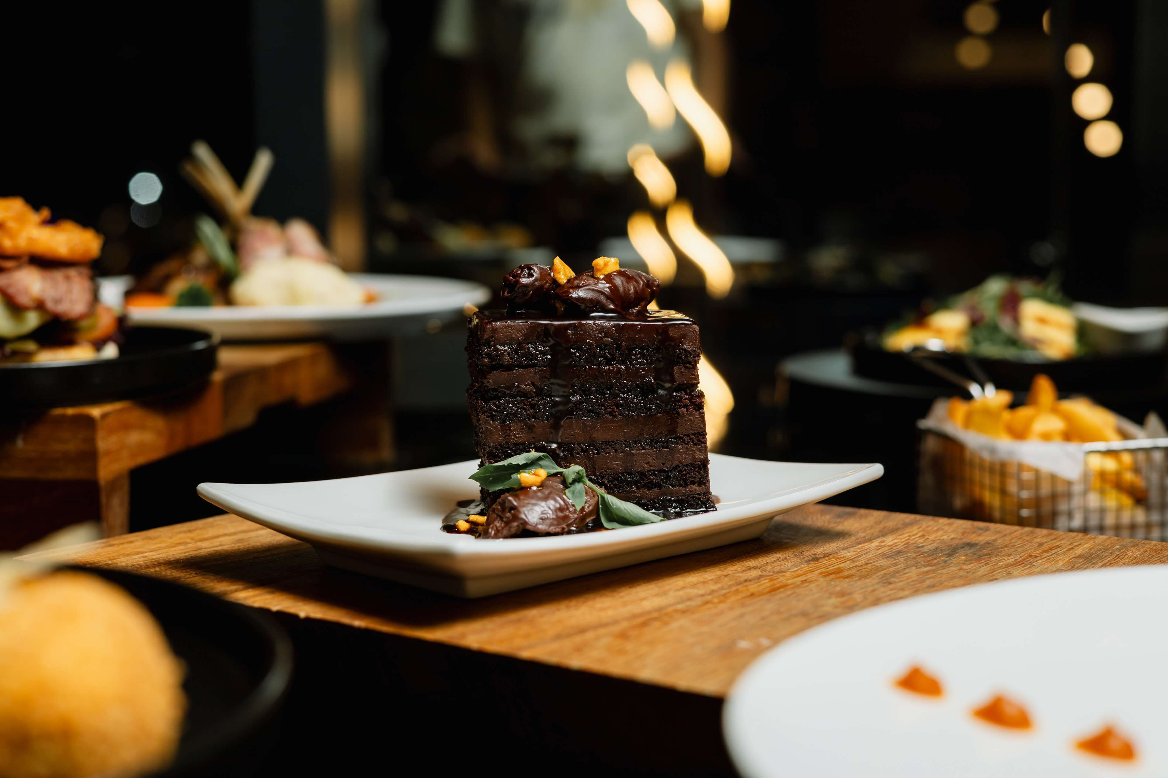 A dessert plate with a chocolate cake, garnished with nuts and leaves, on a white square plate.