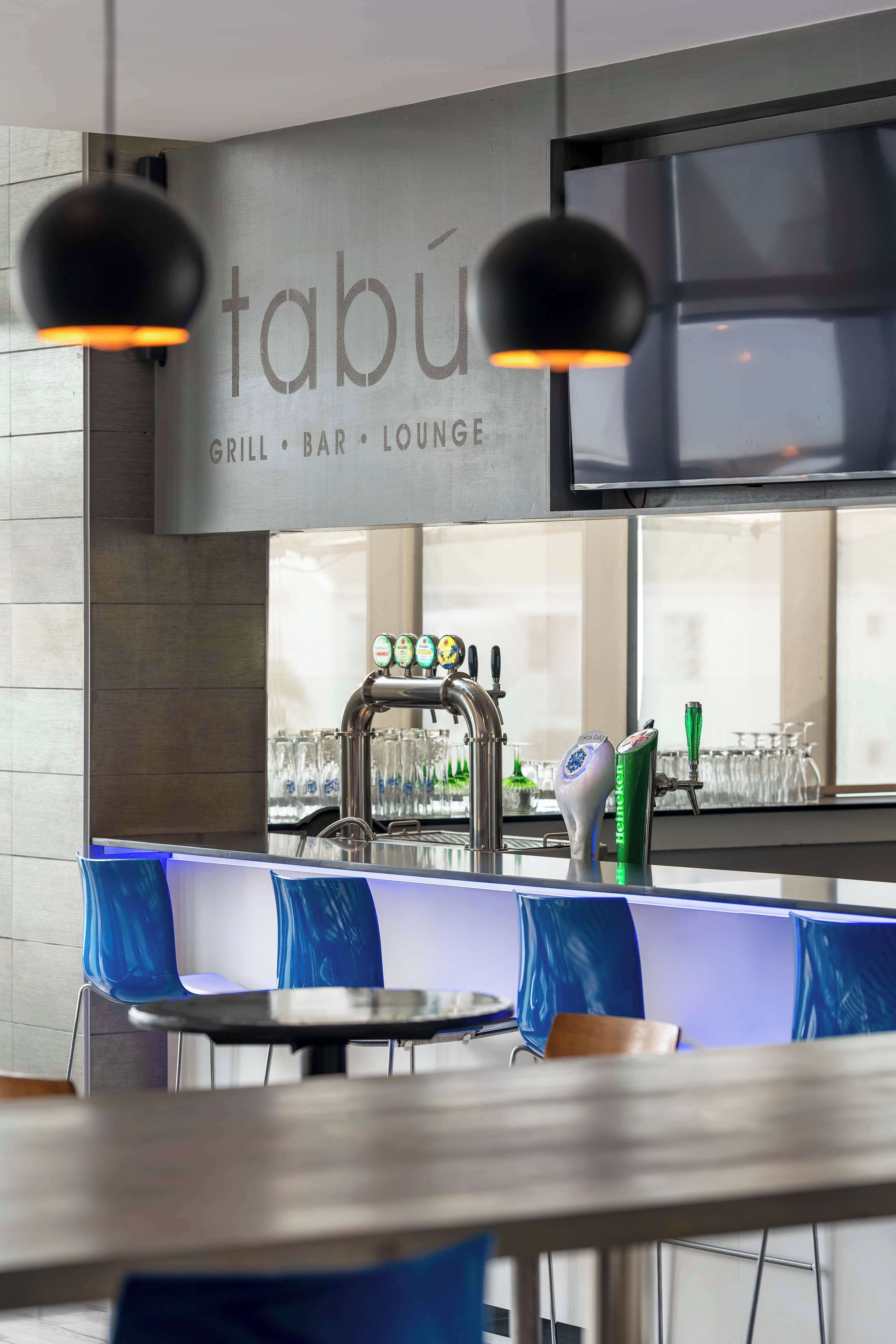 A modern bar area with a sign reading "tabu GRILL BAR LOUNGE," a tap with multiple beer options, and blue chairs around tables.