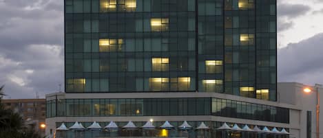 A modern hotel with a glass facade, multiple floors, and a covered entrance area with umbrellas.