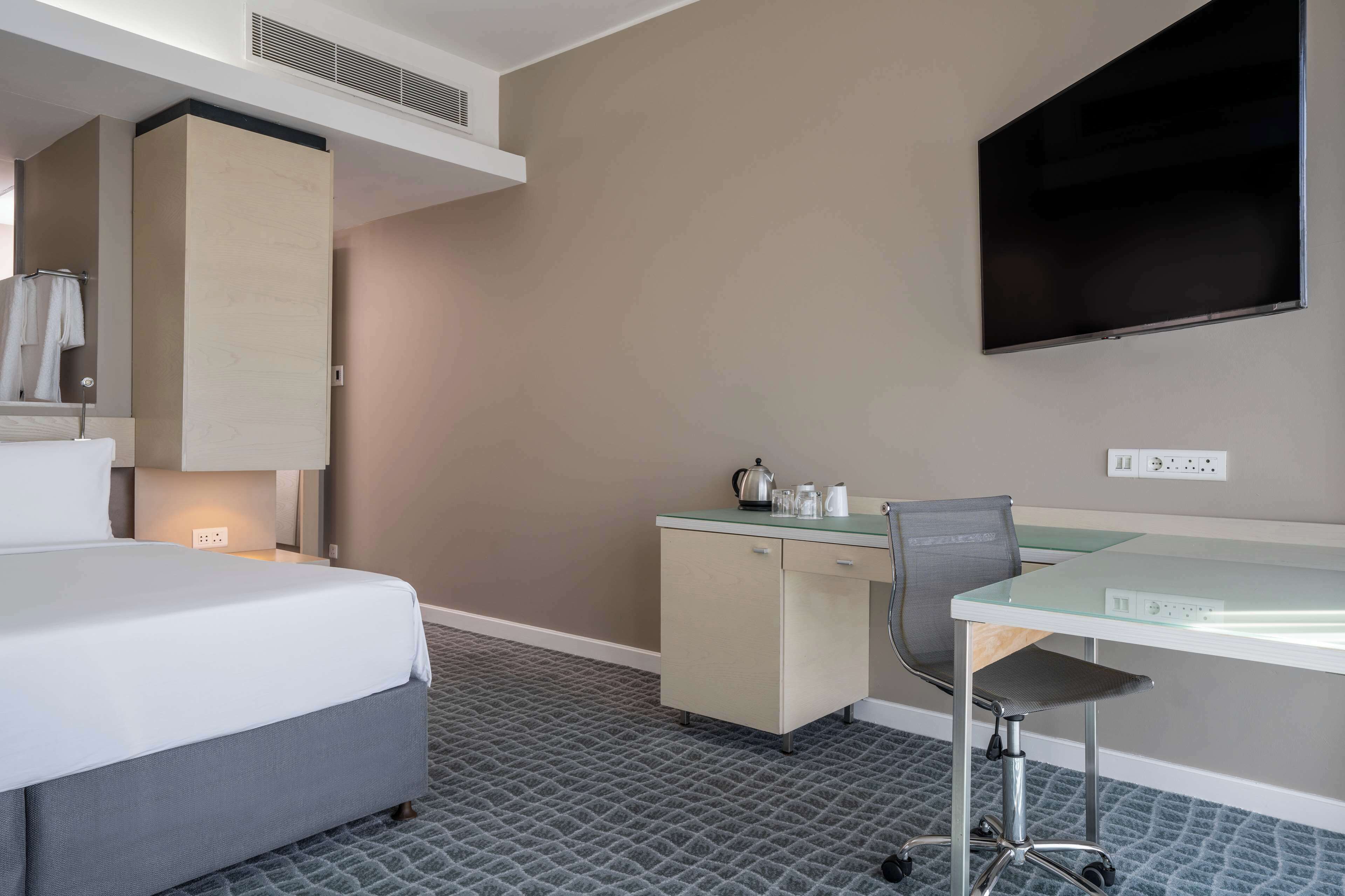 A hotel room with a bed, a desk, a chair, a flat-screen TV, a small kitchenette, and a patterned carpet.