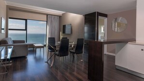 Une chambre d’hôtel moderne dotée d’une grande fenêtre donnant sur la mer, d’un coin salon avec un canapé et des fauteuils, d’une table à manger et d’un petit coin cuisine.