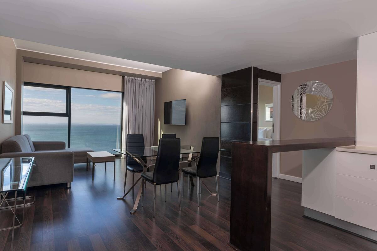 Ein modernes Hotelzimmer mit großem Fenster und Meerblick, einer Sitzecke mit Sofa und Sesseln, einem Esstisch und einer kleinen Küchenzeile.