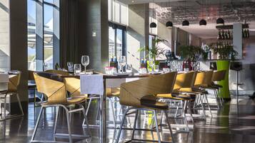 Um restaurante moderno com mesas altas, cadeiras de vime e um bar com garrafas e copos.