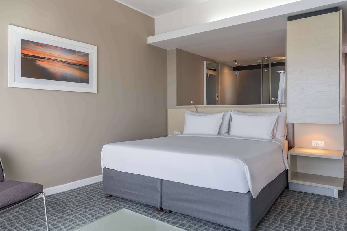 Ein modernes Hotelzimmer mit einem großen Bett, einem Nachttisch, einem Sessel, einem Spiegel und einem gerahmten Bild an der Wand.