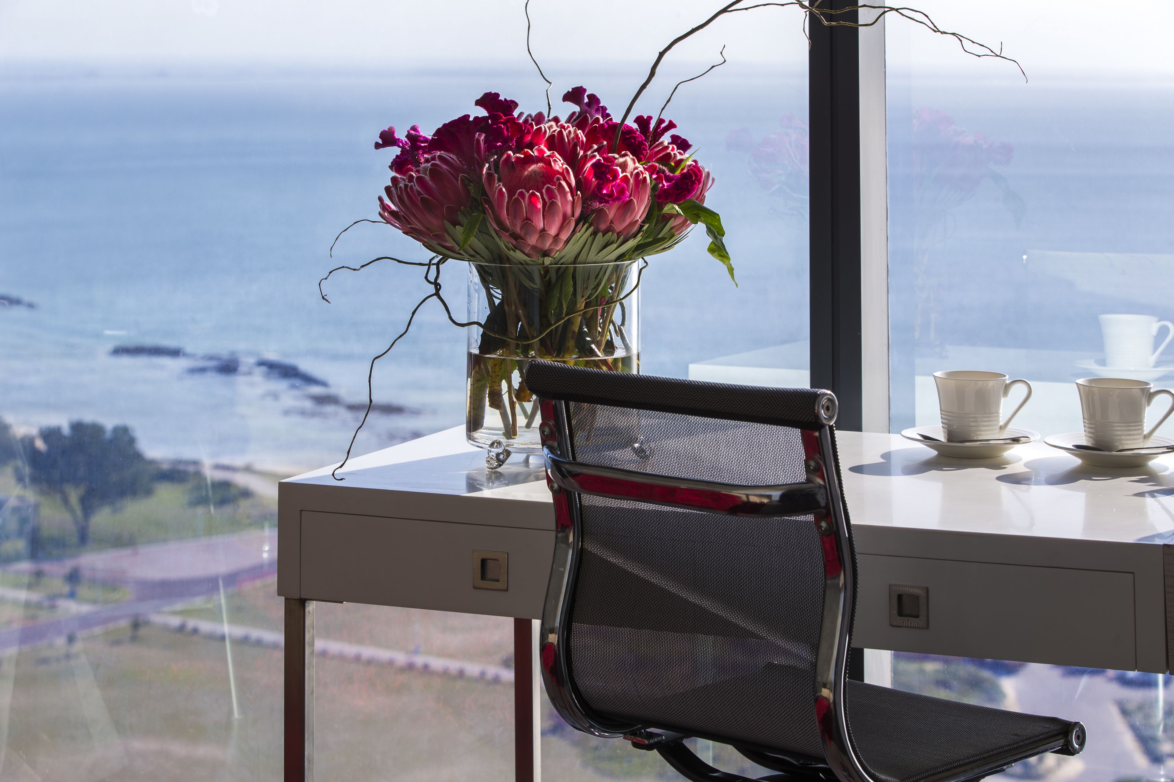 A desk with a vase of flowers, a chair, and a view of the ocean.