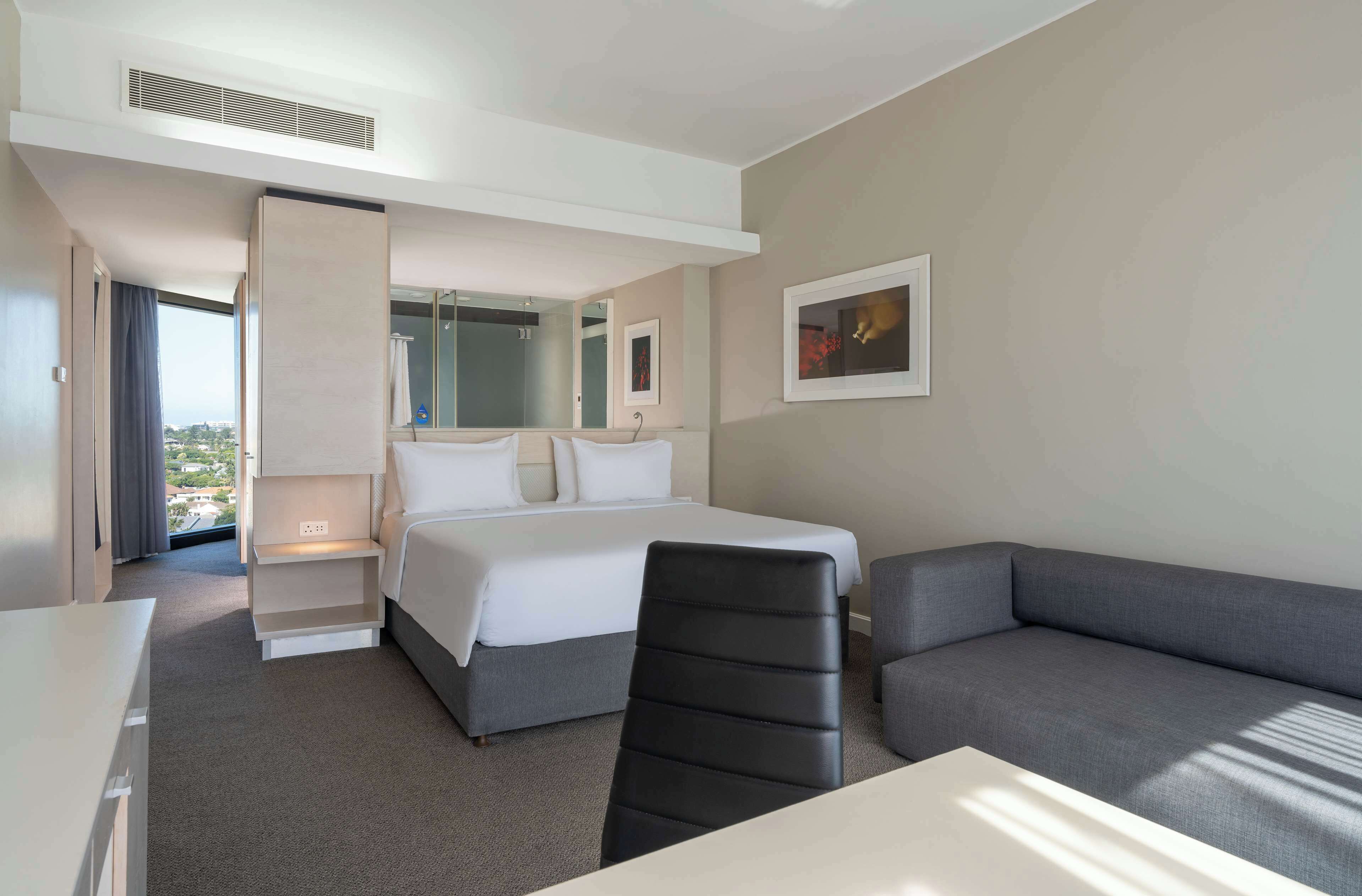A hotel room with a bed, a sofa, a desk, and a chair. There is a window with a view of buildings outside.