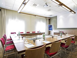 A conference room with a long table, red chairs, a projector screen, and a large window with curtains.