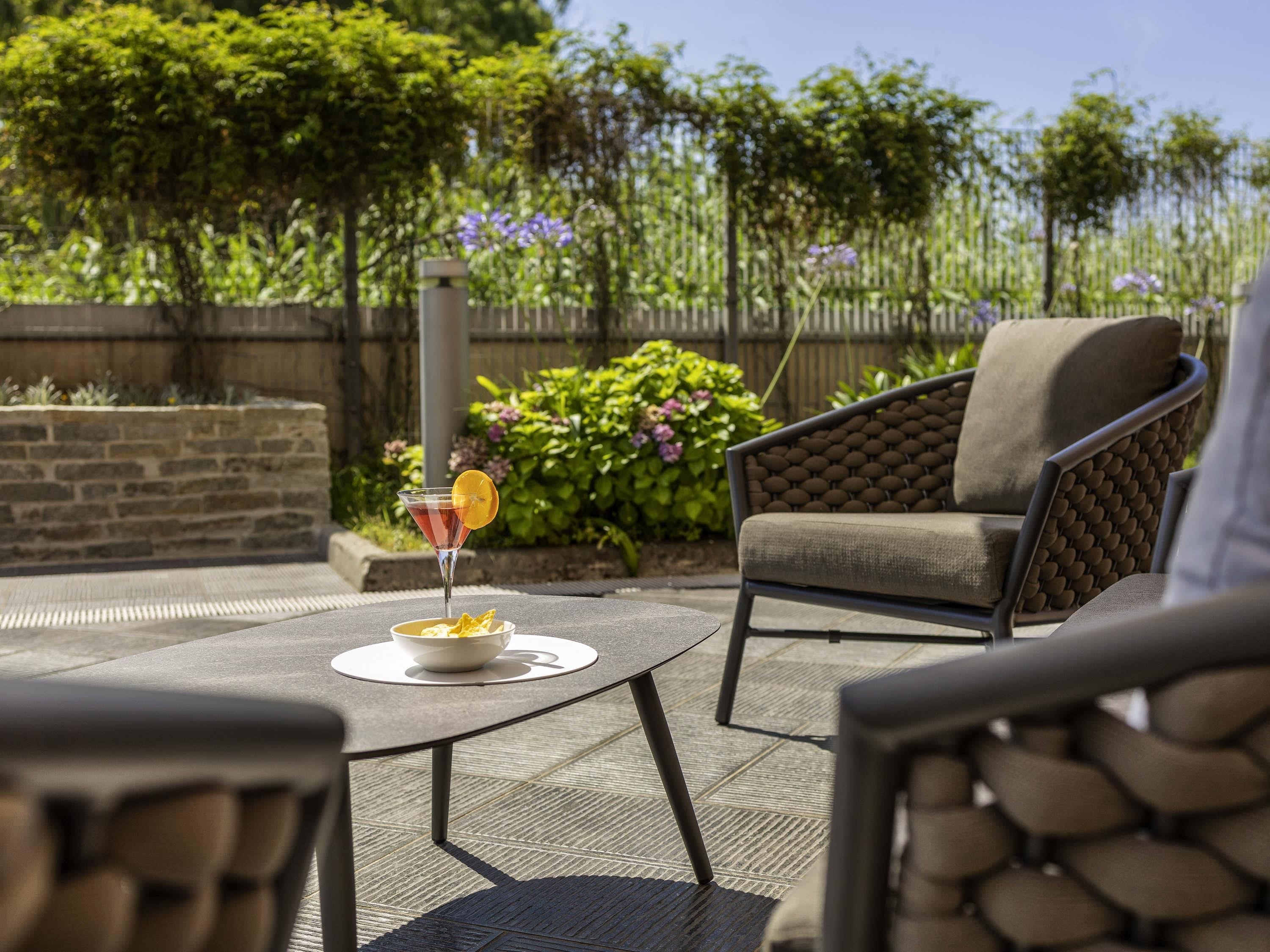 Un patio al aire libre con una mesa redonda, dos sillas, una bebida con una rodaja de naranja y un cuenco de fruta.