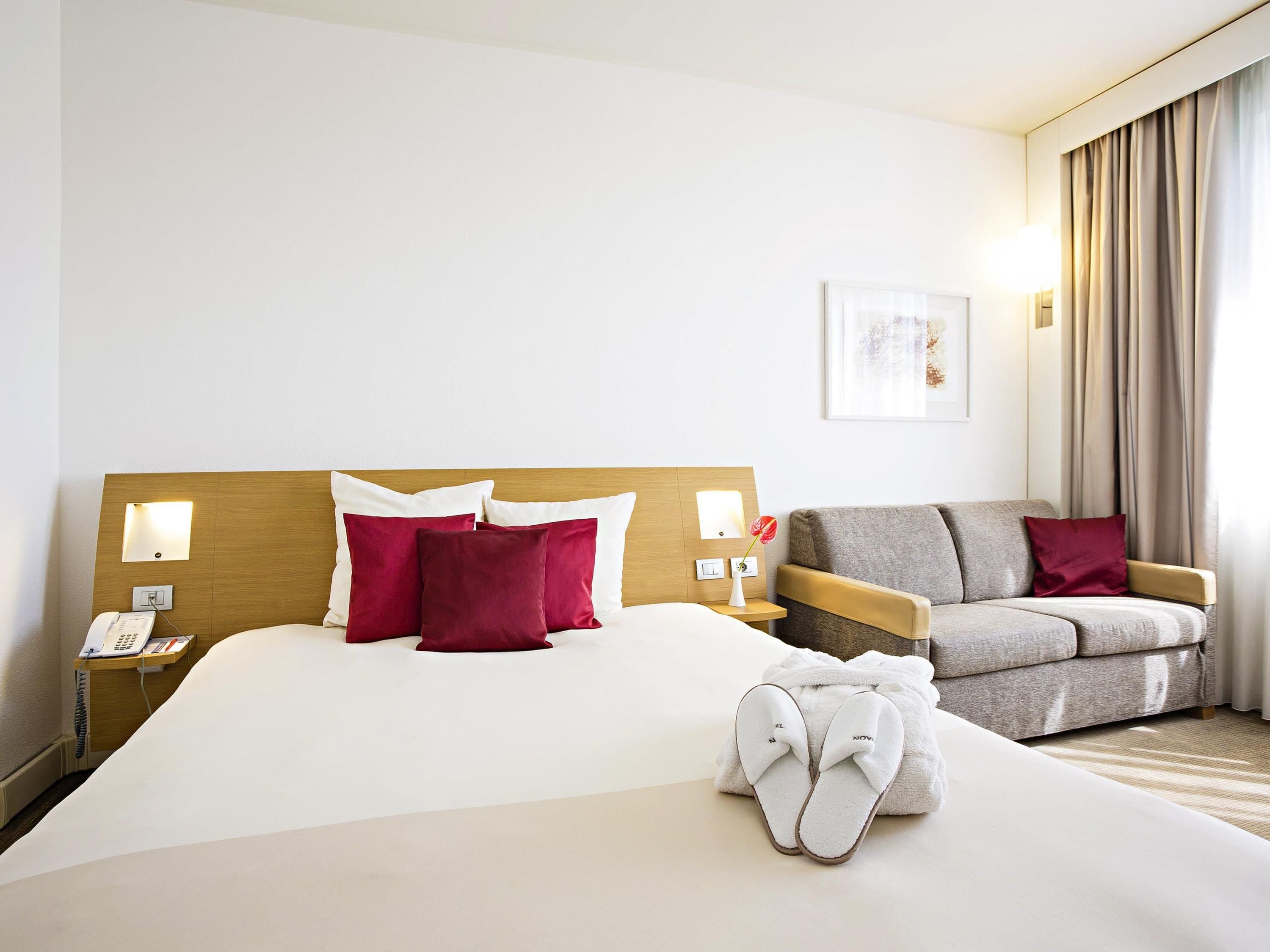 A hotel room with a large bed, a bedside phone, a lamp, a chair, and a pair of slippers on the bed.
