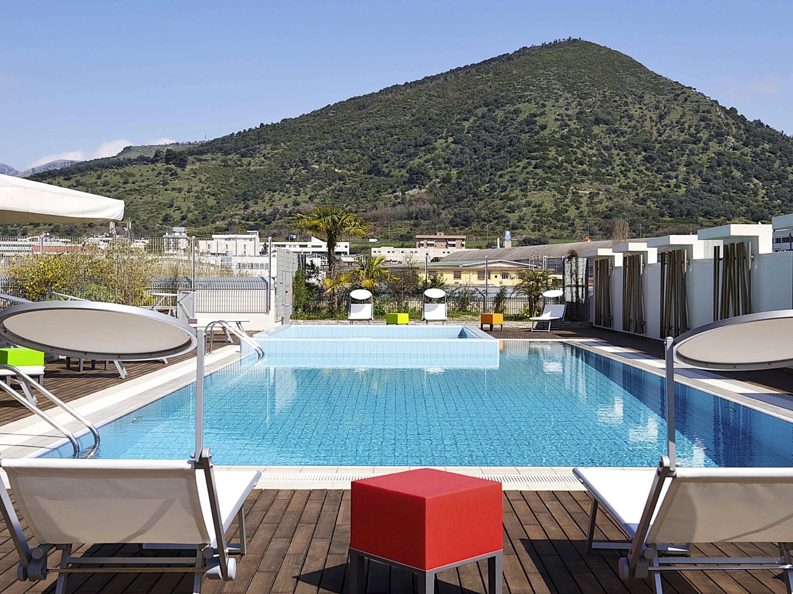 A rooftop pool with a view of a mountain, surrounded by lounge chairs and a red table.