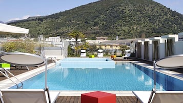 A rooftop pool with a view of a mountain, surrounded by lounge chairs and a red table.