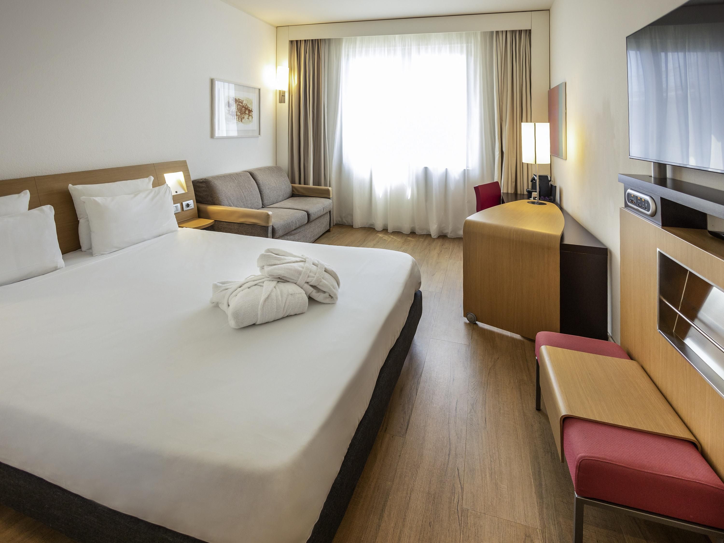 Ein Hotelzimmer mit einem großen Bett, einem Schreibtisch, einem Sessel, einem Fernseher und einem Fenster mit Vorhängen.