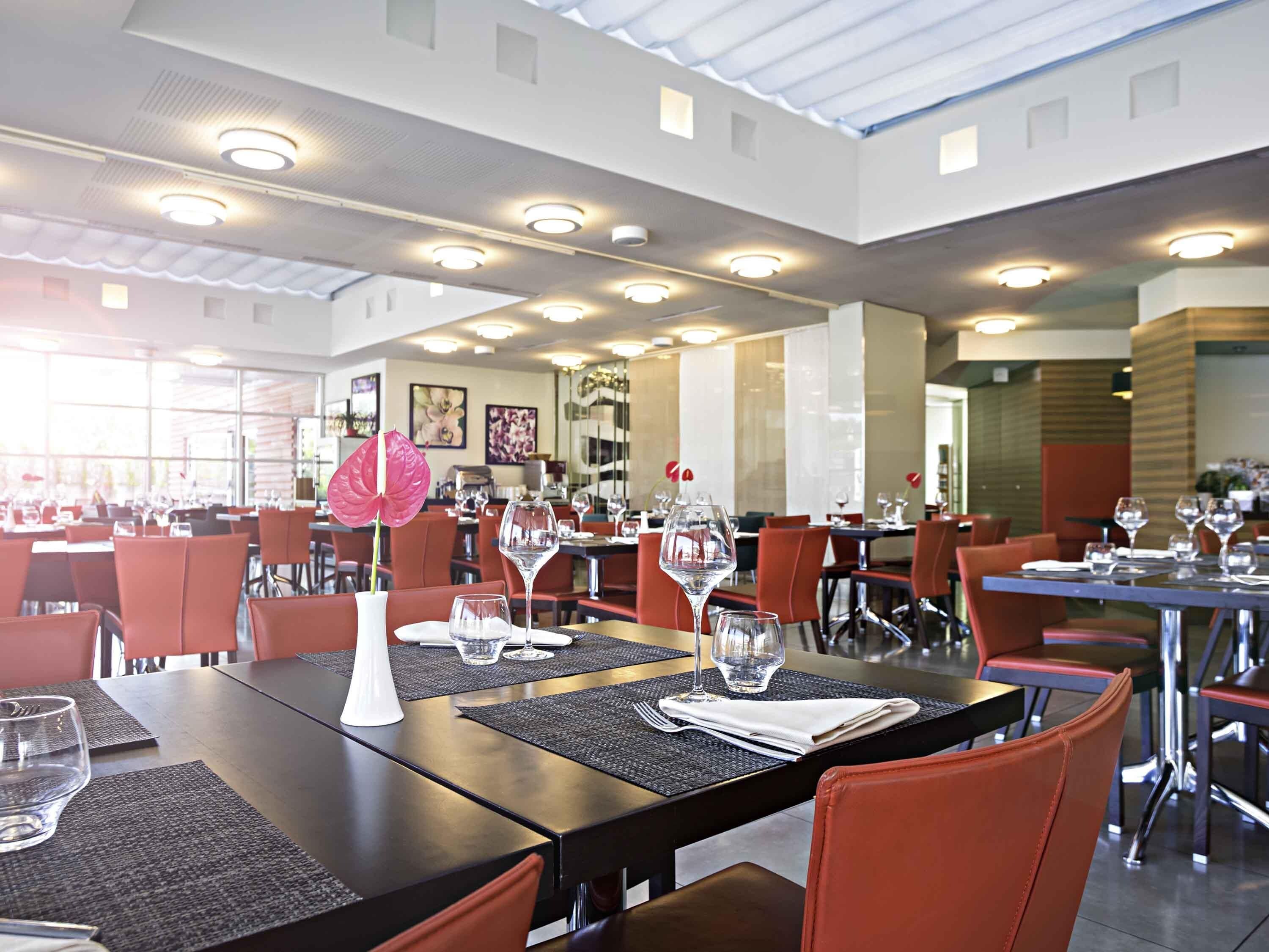 A restaurant with tables set for dining, featuring a modern interior with red chairs and a central decorative plant.