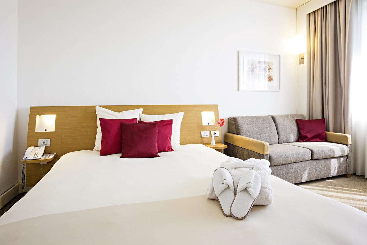 A hotel room with a large bed, a bedside phone, a lamp, a chair, and a pair of slippers on the bed.