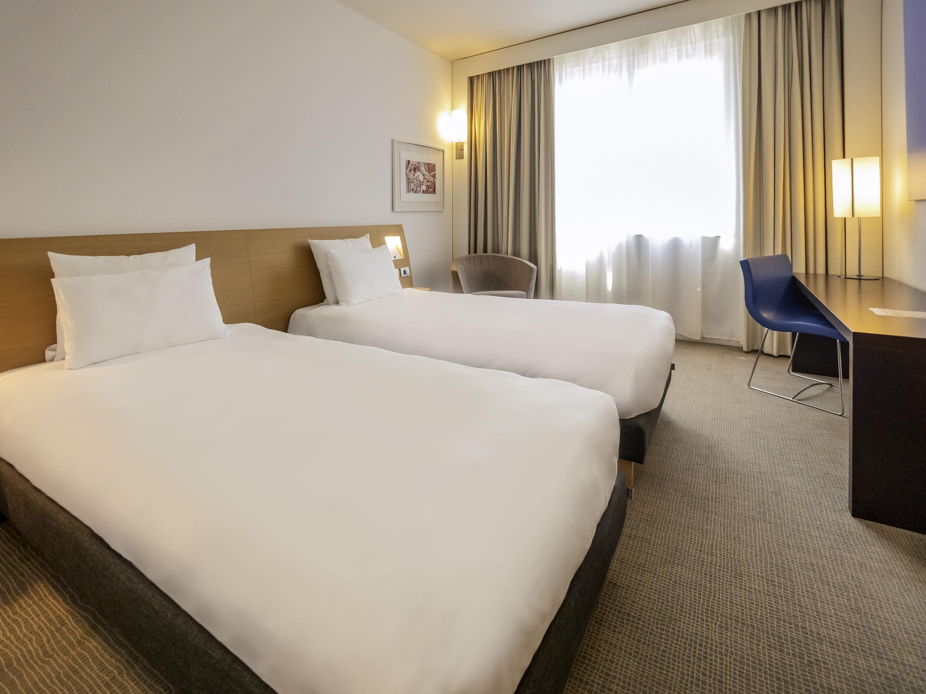 A hotel room with two beds, a desk, a chair, and a large window with curtains.