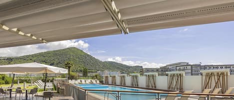Un área de piscina en la azotea con sillones de descanso y mesas, piscina y vistas a la montaña.