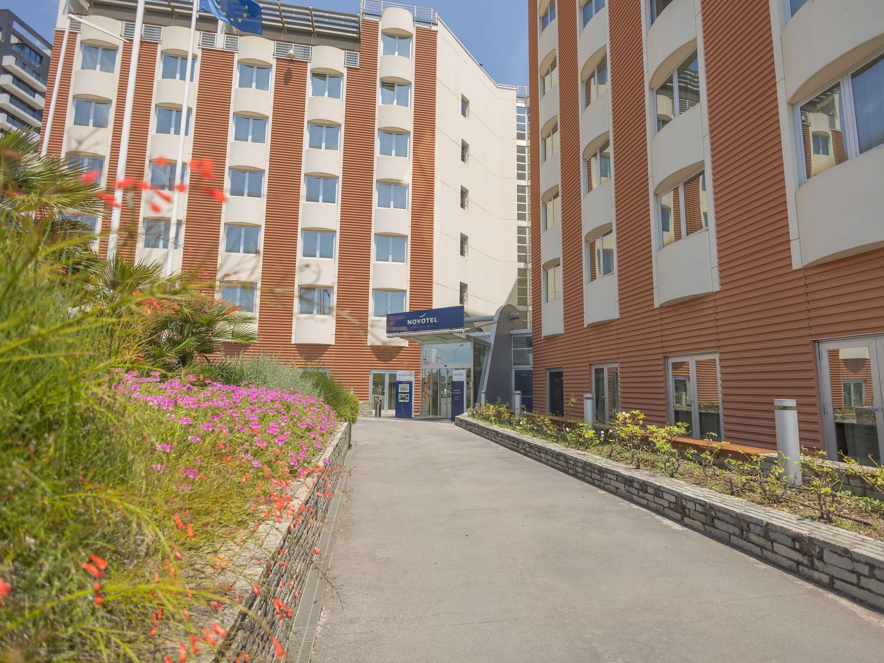 Un hotel dal design moderno, con ingresso centrale, portico coperto e giardino curato con fiori.