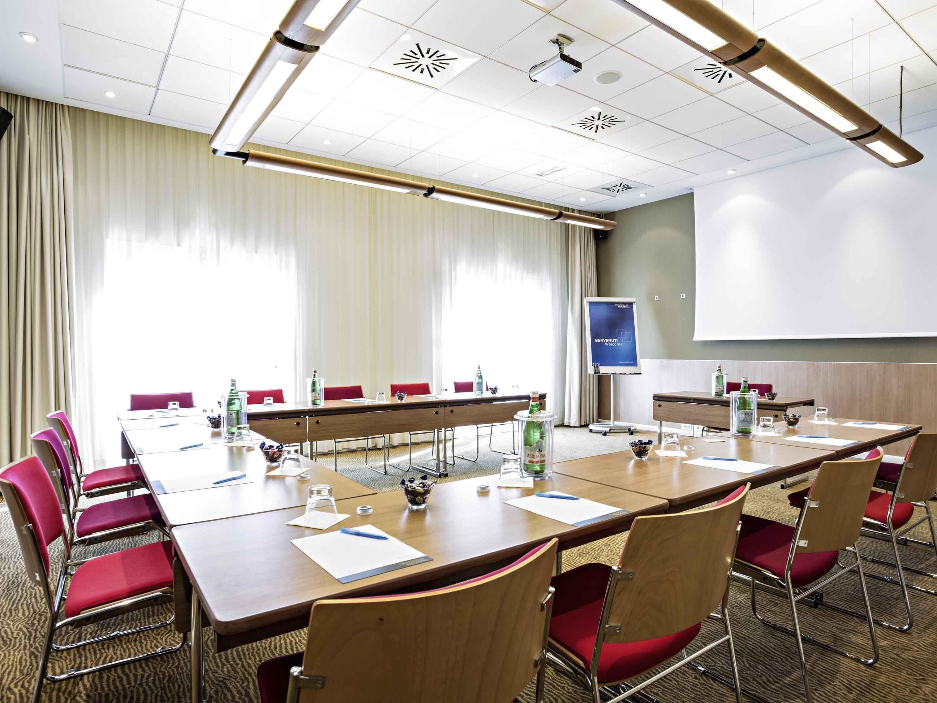 Una sala conferenze con un lungo tavolo, sedie rosse, uno schermo per proiettore e un'ampia finestra con tende.