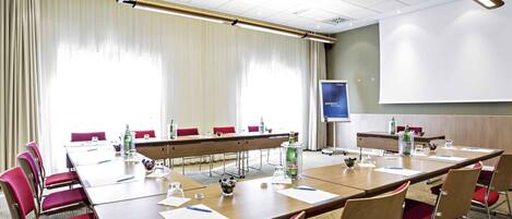 A conference room with a long table, red chairs, a projector screen, and a large window with curtains.