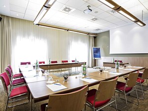 A conference room with a long table, red chairs, a projector screen, and a large window with curtains.